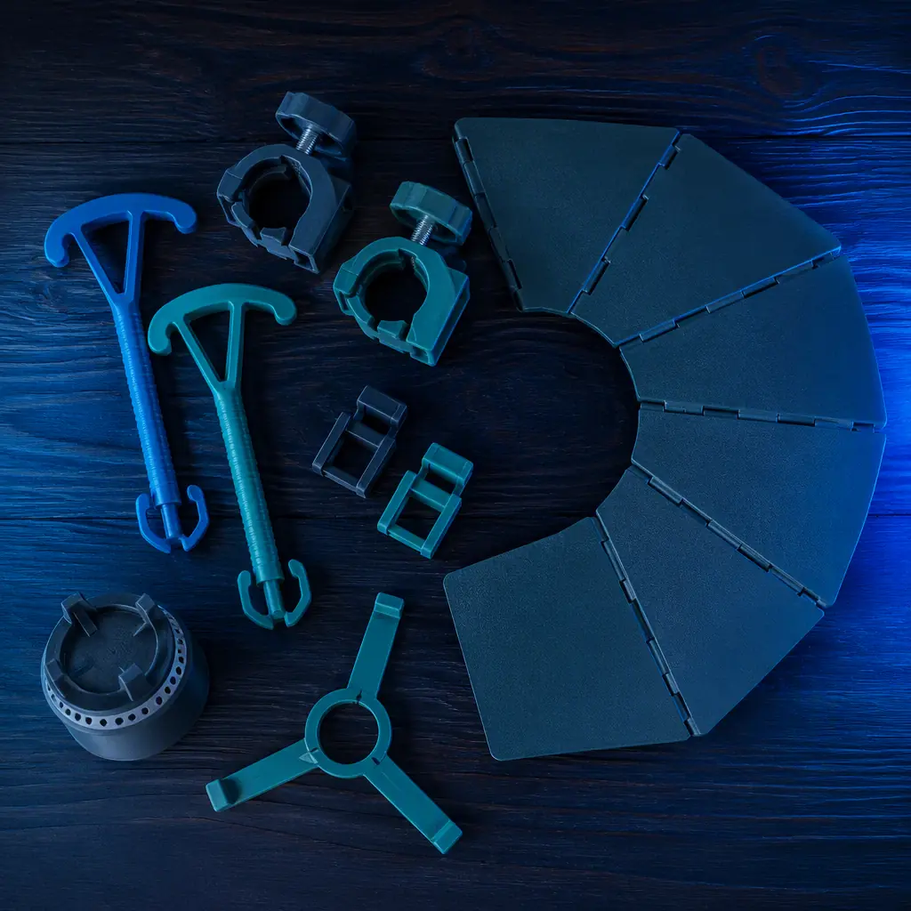3D printed camping accessories arranged on a wooden picnic table: tent stake pullers, lantern mounts, stove windshields and buckles, outdoor soft lighting, photorealistic, no people, no text, no logos